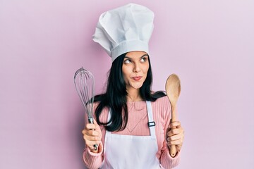 Young hispanic woman wearing baker uniform holding spoon and whisk smiling looking to the side and...