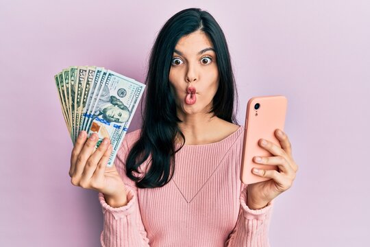 Young hispanic woman using smartphone holding united states dollar banknotes making fish face with mouth and squinting eyes, crazy and comical.