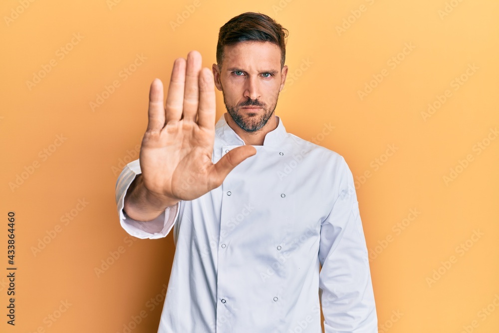 Wall mural Handsome man with beard wearing professional cook uniform doing stop sing with palm of the hand. warning expression with negative and serious gesture on the face.