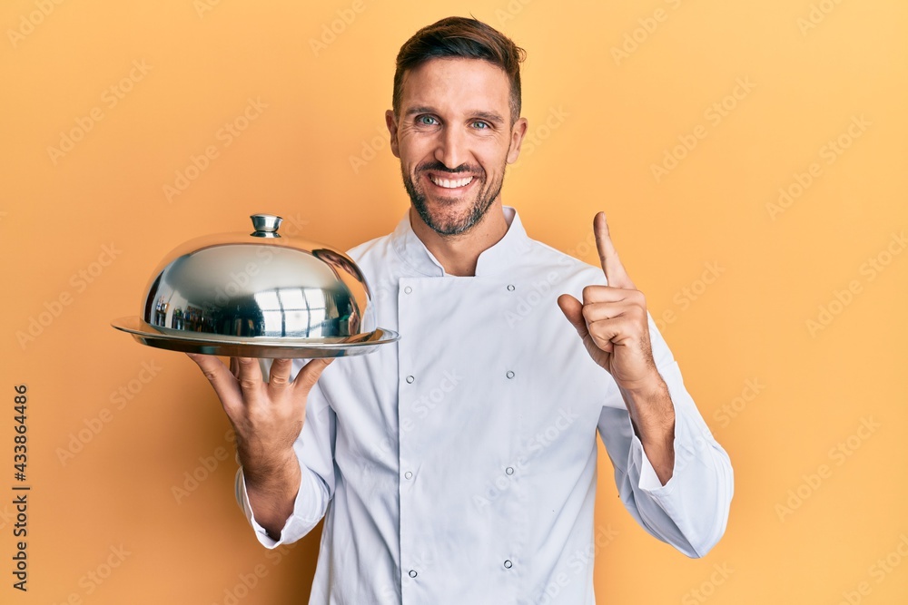 Wall mural Handsome man with beard wearing chef uniform holding silver tray smiling with an idea or question pointing finger with happy face, number one