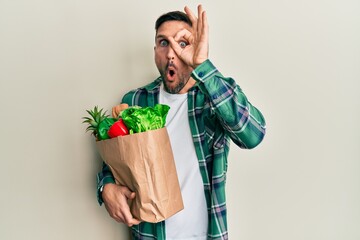 Handsome man with beard holding paper bag with groceries doing ok gesture shocked with surprised...