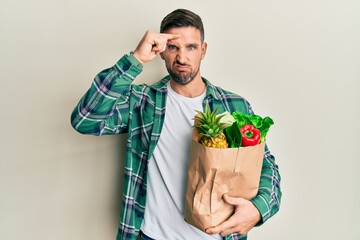 Handsome man with beard holding paper bag with groceries worried and stressed about a problem with hand on forehead, nervous and anxious for crisis