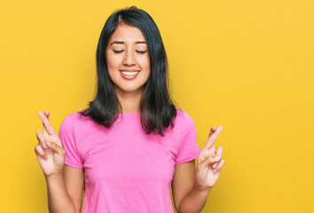 Beautiful asian young woman wearing casual pink t shirt gesturing finger crossed smiling with hope and eyes closed. luck and superstitious concept.