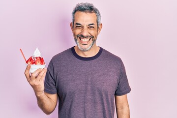 Handsome middle age man with grey hair eating strawberry ice cream looking positive and happy standing and smiling with a confident smile showing teeth