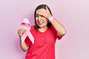 Middle age hispanic woman holding pink cancer ribbon stressed and frustrated with hand on head, surprised and angry face