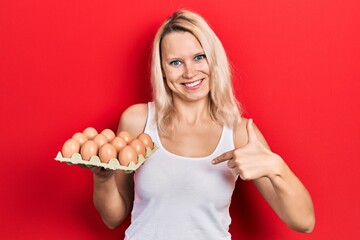Beautiful caucasian blonde woman holding tray of fresh eggs pointing finger to one self smiling happy and proud