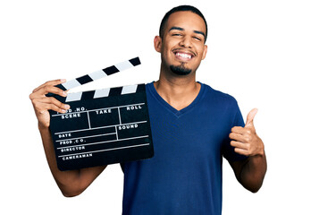 Young african american man holding video film clapboard smiling happy and positive, thumb up doing...