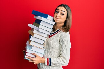 Young brunette student girl holding a pile of books puffing cheeks with funny face. mouth inflated with air, catching air.