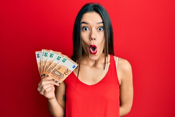 Young hispanic girl holding euro banknotes scared and amazed with open mouth for surprise,...