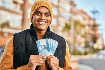 Young latin man smiling happy holding chile pesos banknotes at the city - obrazy, fototapety, plakaty