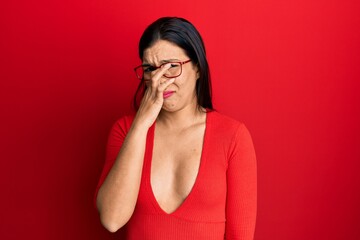 Young latin woman wearing casual clothes and glasses smelling something stinky and disgusting, intolerable smell, holding breath with fingers on nose. bad smell