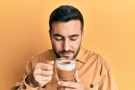 Handsome hispanic man enjoying a cup of coffee over yellow background