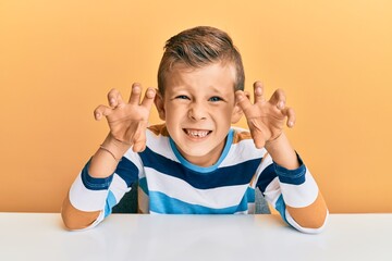 Adorable caucasian kid wearing casual clothes sitting on the table smiling funny doing claw gesture...
