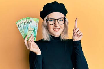 Young blonde girl holding russian 200 ruble banknotes smiling with an idea or question pointing finger with happy face, number one