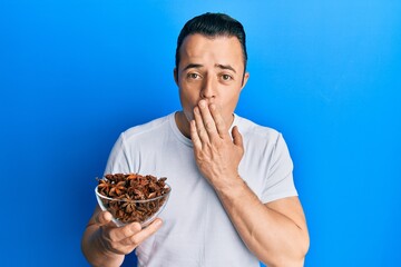 Handsome young man holding bowl of star anise covering mouth with hand, shocked and afraid for...