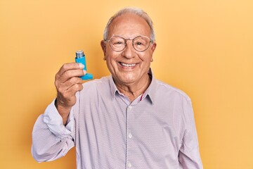 Senior man with grey hair holding medical asthma inhaler looking positive and happy standing and...