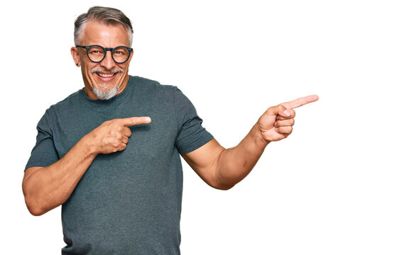 Middle age grey-haired man wearing casual clothes and glasses smiling and looking at the camera pointing with two hands and fingers to the side.