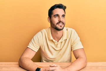 Young hispanic man wearing casual clothes sitting on the table looking away to side with smile on face, natural expression. laughing confident.