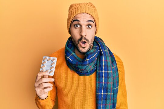 Young hispanic man wearing wool sweater and winter scarf holding pills scared and amazed with open mouth for surprise, disbelief face
