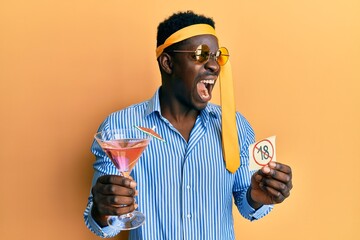 Handsome black man drunk wearing tie over head drinking cocktail holding under 18 sign angry and...