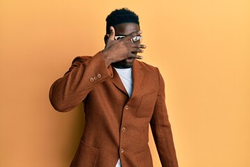 Handsome black man wearing elegant clothes and glasses peeking in shock covering face and eyes with hand, looking through fingers afraid
