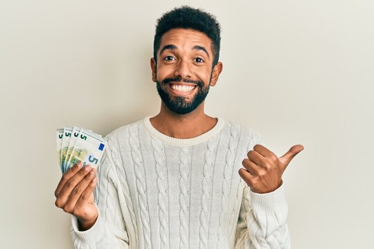 Handsome hispanic man with beard holding bunch of 5 euro banknotes pointing thumb up to the side smiling happy with open mouth