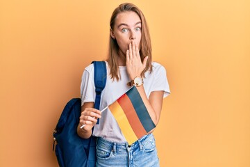 Beautiful blonde woman exchange student holding germany flag covering mouth with hand, shocked and...