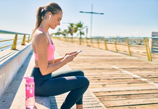 Young blonde sportswoman doing exercise using smartphone sitting on the bench at the promenade.
