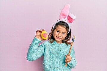 Little beautiful girl wearing cute easter bunny ears holding colored egg smiling with a happy and cool smile on face. showing teeth.