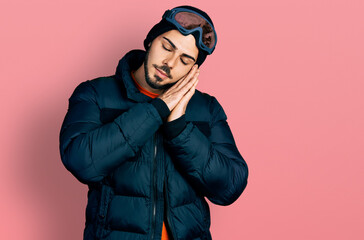 Young hispanic man with beard wearing snow wear and sky glasses sleeping tired dreaming and posing with hands together while smiling with closed eyes.