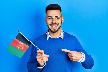 Young hispanic man with beard holding afghanistan flag smiling happy pointing with hand and finger