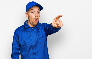 Bald man with beard wearing builder jumpsuit uniform pointing with finger surprised ahead, open mouth amazed expression, something on the front