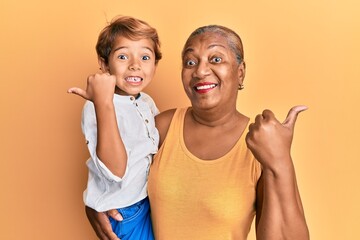 Hispanic grandson and grandmother together over yellow background pointing thumb up to the side smiling happy with open mouth