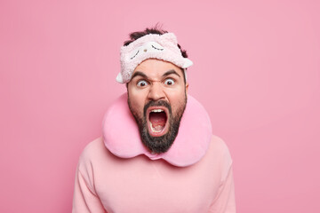 Outraged bearded man screams louldly keeps mouth widely opened expresses negative emotions being angry with someone who interrupts sleep wears blindfold travel pillow around neck isolated on pink wall