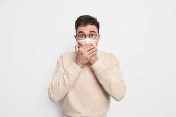 Protection against disease. Shocked frightened man keeps hands on mouth wears protective face mask to prevent coronavirus or contagious disease poses against white background stunned by new reality