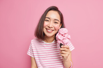 Lovely dark haired Asian woman enjoys eating tasty strawberry ice cream during hot summer day has upbeat mood pleased face expression dressed casually poses with favorite frozen dessert indoor