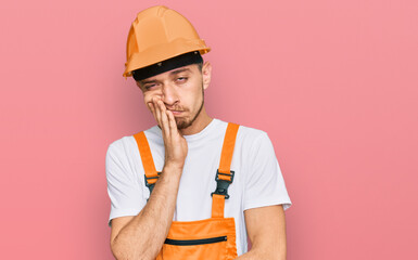 Hispanic young man wearing handyman uniform and safety hardhat thinking looking tired and bored with depression problems with crossed arms.