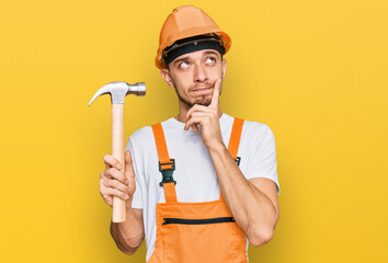 Hispanic young man wearing hardhat holding hammer serious face thinking about question with hand on chin, thoughtful about confusing idea