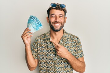 Handsome man with beard holding 100 brazilian real banknotes smiling happy pointing with hand and...