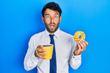 Handsome business man with beard eating doughnut and drinking coffee in shock face, looking skeptical and sarcastic, surprised with open mouth