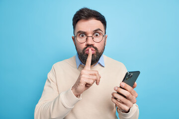 Surprised mysterious man makes silence gesture tells secret information uses mobile phone for online chatting and distance working wears spectacles casual sweater isolated over blue background