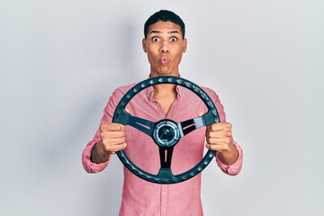 Young african american guy holding steering wheel making fish face with mouth and squinting eyes, crazy and comical.