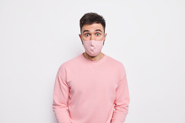 Prevention and safety concept. Surprised young man wears protective mask on face prevents coronavirus spread finds out shocking statistics dressed in pink sweater isolated over white background