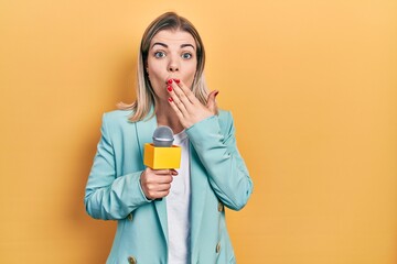 Beautiful caucasian woman holding reporter microphone covering mouth with hand, shocked and afraid...