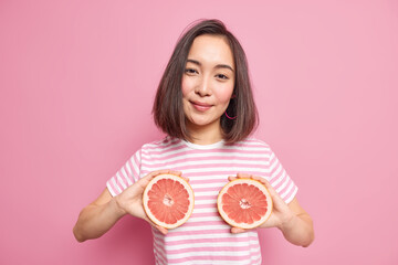 Pretty brunette woman with dark hair holds two halves of grapefruit over body dressed in casual striped t shirt looks satisfied at camera isolated over pink background. Female holds juicy fruit