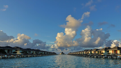 Obraz premium Morning in the Maldives. There are picturesque golden clouds in the azure sky. Above the aquamarine ocean, there are two rows of water villas. Reflection on the surface of the water. No people