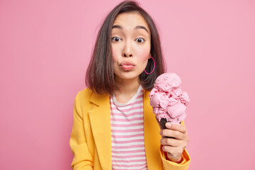 Pretty millennial Asian woman keeps lips folded holds appetizing ice cream in black waffle spends day off walking outside enjoys summer time eats tasty frozen cold dessert wears yellow jacket