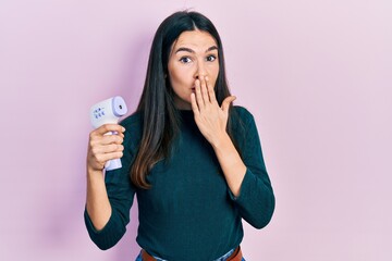 Young brunette woman holding thermometer covering mouth with hand, shocked and afraid for mistake....