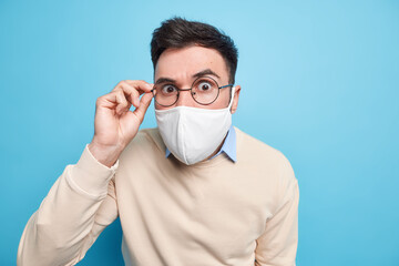 Protection against coronavirus. Serious young man uses disposable face mask in public place during quarantine keeps hand on rim of spectacles dressed in sweater isolated over blue background