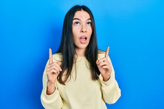Young brunette woman wearing casual sweatshirt amazed and surprised looking up and pointing with fingers and raised arms.
